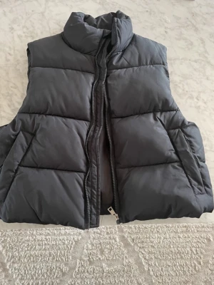 Svart dunväst från 157, storlek S - Snygg svart dunväst från 157 med hög krage och dragkedja framtill. Västen har en puffig design och är perfekt för lager på lager. Tillverkad i ett mjukt och slitstarkt material som håller dig varm under kyliga dagar.