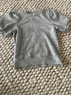 Grå kortärmad topp med puffärm - En stilren grå topp med korta puffärmar och rund hals. Toppen har en mjuk och lite tjockare struktur, perfekt för en avslappnad men ändå trendig look. Passar till jeans eller kjol för en enkel outfit.