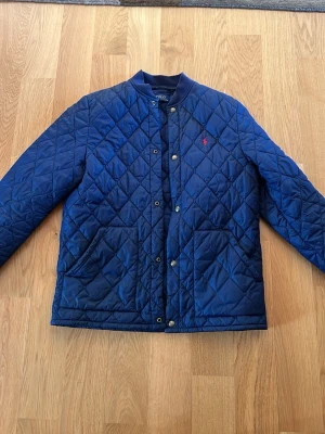 Blå quiltad dunjacka från Polo Ralph Lauren - Snygg blå dunjacka från Polo Ralph Lauren med quiltat mönster, tryckknappar framtill och två stora fickor. Jackan har en liten röd logga broderad på bröstet och ribbade muddar vid ärmsluten. Perfekt för kalla vinterdagar.