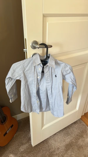 Ljusblå skjorta från Ralph Lauren - Klassisk ljusblå skjorta från Ralph Lauren med lång ärm och knappar framtill. Skjortan har en broderad marinblå logga på bröstet och är tillverkad i mjuk bomull. Perfekt för en clean och stilren look.