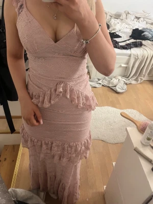 Ljusrosa spetsklänning med volanger - Romantisk långklänning i ljusrosa spets med v-ringning och breda axelband. Klänningen har flera volangdetaljer längs midjan och nederdelen, samt figurnära passform. Perfekt för sommarens alla tillfällen.