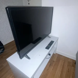 Platt-TV med fjärrkontroll - Stilren platt-TV med tillhörande fjärrkontroll. Skärmen ser ut att vara i gott skick utan synliga repor eller skador. Perfekt för vardagsrummet eller sovrummet. Står stadigt på två ben och levereras med fjärrkontroll för enkel användning. 36 tum