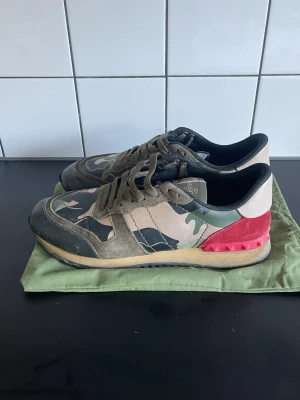 Valentino rockrunners - Snygga sneakers från Valentino Garavani med camomönster i grönt, beige och svart. Skorna har detaljer i mocka, läder och textil samt en röd hälpanel med nitar. Klassisk låg modell med gröna skosnören och svart innersula. Allt på sista bilden tillkommer. Lite små sliten och tidigare håll på främre delen av skon (bara högerfoten) hör av er om ni har några frågor!!!!