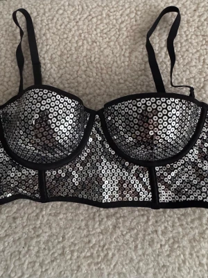 Svart bh med paljetter och justerbara band - Snygg svart bh med glittriga paljetter framtill och justerbara axelband. Bh:n har hakknäppning bak och är tillverkad i ett glansigt material som ger en lyxig känsla. Perfekt för dig som vill ha något extra till outfiten. Använd en gång. Storleklapp avklippt.