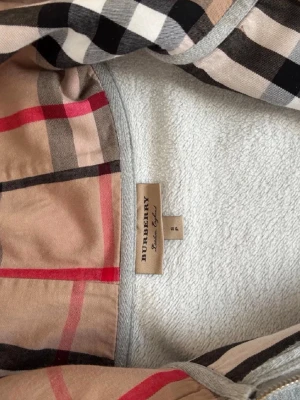 hoodie Burberry med dragkedja (BUD:600kr) - Snygg grå hoodie från Burberry med dragkedja och klassiskt rutigt foder i huvan. Hoodien har två fickor framtill, justerbar huva med snören och ribbade muddar. Perfekt för en chill och stilren look. Materialet är mjukt och bekvämt, troligen bomullsblandning.