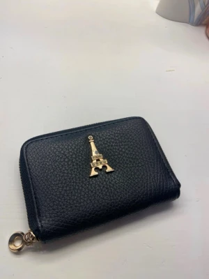 Svart plånbok med guldiga detaljer  - Snygg svart plånbok i konstläder med guldfärgad dragkedja och en dekorativ Eiffeltornet-detalj på framsidan. Praktisk storlek som får plats i fickan eller väskan. Perfekt för kort och kontanter.