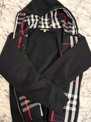 Burberry hoodie - Svart hoodie från Burberry med dragkedja och huva. Insidan av huvan har ett klassiskt rutigt mönster i svart, vitt och rött. Tröjan har långa ärmar, ribbade muddar och mjukt fleecefoder. Perfekt för dig som gillar stilren streetwear med en twist. Passar både m/s. Dragkedjan är trasig men den är nice ändå 