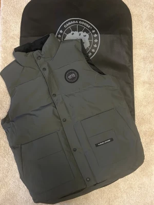 Grå dunväst från Canada Goose - Snygg dunväst från Canada Goose med hög krage och två stora framfickor. Västen har ett svart Canada Goose-märke på bröstet och stängs med tryckknappar. Perfekt för lager-på-lager och riktigt varm tack vare dunfyllningen.