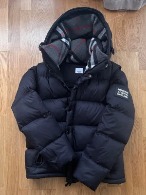 Svart pufferjacka från Burberry - Snygg svart pufferjacka från Burberry med huva och patch på ärmen där det står 'Burberry London England'. Jackan har en quiltad design och dragkedja framtill. Insidan av huvan har ett klassiskt rutigt mönster i grått, vitt och rött. Perfekt för kalla vinterdagar. Jag har använt den bara 2 månader under vintern annars den är nästan ny. 
