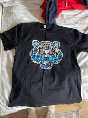  Svart Kenzo t-shirt med tigertryck - Säljer en svart t-shirt från Kenzo med det ikoniska tigerhuvudet i vitt, blått och grått på bröstet. T-shirten är i 100% bomull och har en klassisk passform med korta ärmar och rund hals. Perfekt statement-plagg för dig som gillar streetwear.