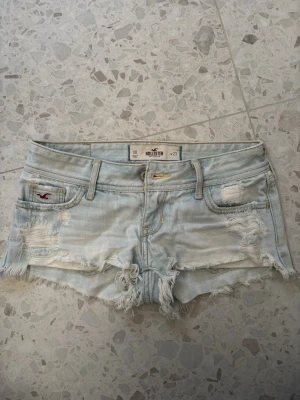 Hollister jeansshorts - Säljer ett par ljusblå jeansshorts från Hollister med slitna detaljer. Shortsen har låg midja och är gjorda i bomull. Perfekta för sommaren och ger en avslappnad vibe. Midjemåttet 36-37cm