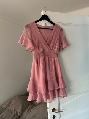 Rosa volangklänning med v-ringning - Supersöt rosa klänning med v-ringning, korta volangärmar och smock i midjan. Kjolen har dubbla volanglager. Materialet ser ut att vara lätt och luftigt, troligen polyester eller viskos. St 34. Skriv om ni har några frågor!