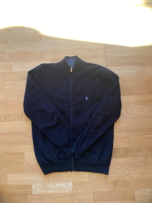 Mörkblå ziptröja från Polo Ralph Lauren - Mörkblå ziptröja från Polo Ralph Lauren med ståkrage och dragkedja hela vägen. Tröjan har det klassiska broderade logot på bröstet och ribbade muddar vid ärmslut och nederkant. Perfekt för lager på lager och stilren look.