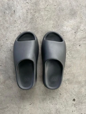 Svarta Adidas tofflor i skum - Säljer ett par svarta Adidas tofflor i mjukt skummaterial. De har en minimalistisk design med öppen tå och räfflad sula för extra grepp. Perfekta för chill dagar hemma eller till stranden. Adidas-logga syns diskret på fotbädden.