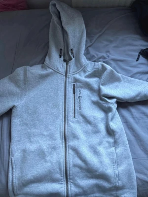 Ljusgrå hoodie från sail racing - Säljer en ljusgrå hoodie från sail racing med dragkedja hela vägen, huva med dragsko och en bröstficka med dragkedja. Hoodien har en clean look och är tillverkad i mjukt sweatshirtmaterial. Perfekt för chill dagar eller när du vill ha en enkel stil.