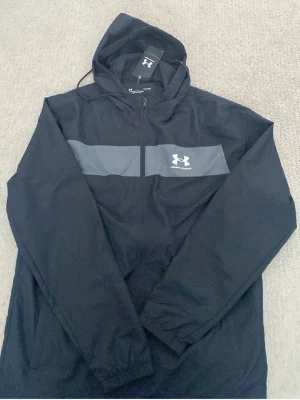 Svart vindjacka från Under Armour - Snygg svart vindjacka från Under Armour med huva och dragkedja framtill. Jackan har en grå panel över bröstet med logga och är tillverkad i lätt, vindtätt material. Perfekt för blåsiga dagar och sportiga outfits. Utan tag!