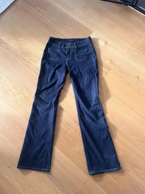 Mörkblå bootcut jeans från MQ - Snygga mörkblå bootcut jeans från MQ med kontrastsömmar och två framfickor med lock och knappdetalj. Klassisk femficksmodell i mjukt denimtyg. Perfekta för en avslappnad men trendig look.