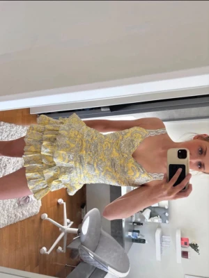 Gul och vit volangklänning - Söt kortklänning med volanger i flera lager, gul och vit med blommigt mönster. Klänningen har smala axelband och v-ringad halsringning. Perfekt för sommarens alla tillfällen.