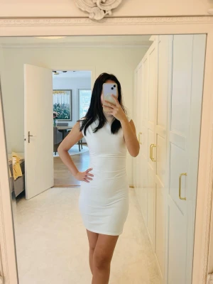 Ny Snygg Vit ärmlös kortklänning från Meshki - Säljer en stilren vit kortklänning med figurnära passform och hög hals. Klänningen är ärmlös och sydd i ett mjukt, stretchigt material som ger en snygg siluett. Perfekt för dig som gillar minimalistisk och modern stil.