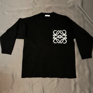 Svart stickad tröja från Loewe - Snygg svart stickad tröja från Loewe med rund halsringning och en stor bröstficka prydd med vitt broderat Loewe-monogram. Tröjan har korta ärmar och en rak passform som ger en clean och modern look.