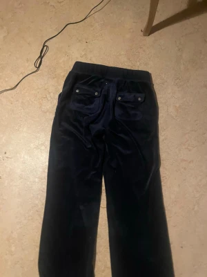Mörkblå velourbyxor med fickor - Säljer ett par mörkblå mjukisbyxor från juicy couture i mjuk velour med resår i midjan och snörning. Byxorna har två stora fickor fram och två bakfickor med knappdetaljer. Perfekta för en chill och avslappnad stil.