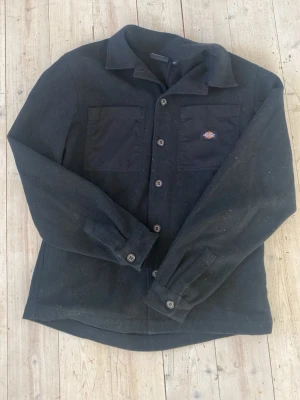 Svart overshirt från Dickies - Stilren svart overshirt från Dickies med klassisk krage och knäppning framtill. Två bröstfickor, varav en med Dickies-logga. Tillverkad i ett robust material som känns slitstarkt och passar perfekt till lager-på-lager. Lång ärm med knapp vid ärmslut.