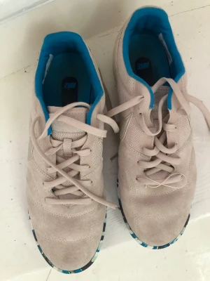 Nike beige sneakers med blå detaljer - Snygga beige sneakers från Nike med mockaliknande material och meshdetaljer. Insidan är klädd i klarblått tyg och yttersulan har ett unikt mönster i blått, svart och vitt. Klassisk snörning och diskret Nike-logga på sidan.