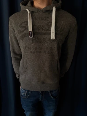 Mörkgrå Superdry hoodie med tryck - Mörkgrå hoodie från Superdry med stort ton-i-ton-tryck på bröstet och vita, breda dragsnören i huvan. Klassisk känguruficka framtill och ribbade muddar vid ärmslut och nederkant. Perfekt för en avslappnad stil. Hör gärna av dig vid minsta fundering.