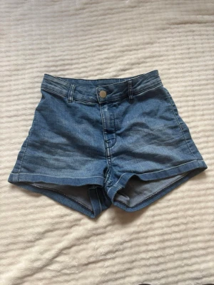 Högmidjade blå jeansshorts - Snygga högmidjade jeansshorts i klassisk blå denim med knapp och dragkedja framtill. Shortsen har två fickor bak och bälteshällor i midjan. Perfekta för varma dagar och enkel att matcha med olika toppar.