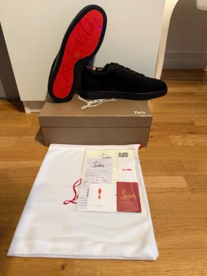 Svarta Louboutin sneakers med röd sula - Snygga svarta sneakers från Christian Louboutin med ikonisk röd sula och diskret logga på sidan. Skorna har en klassisk låg siluett och är tillverkade i mocka med svarta skosnören. Perfekta för dig som vill sticka ut med exklusiv stil.