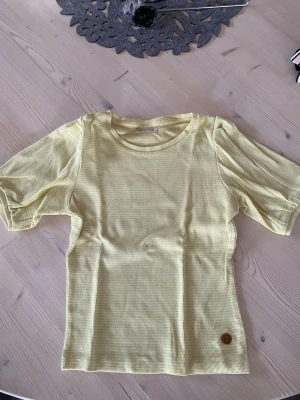 Gulvit randig t-shirt med puffärm - Säljer en söt gulvit randig t-shirt med puffärmar och rund hals. T-shirten har ett litet brunt märke längst ner. Perfekt för dig som gillar en fräsch och lekfull stil. Köparen står för frakt.