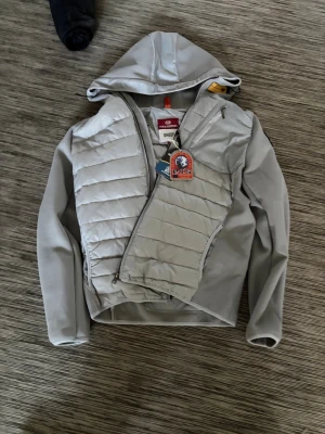 Ljusgrå pufferjacka från Parajumpers - Snygg ljusgrå pufferjacka från Parajumpers, modell Sheen. Jackan har huva, quiltad front och ärmar i stretchigt material. Praktisk bröstficka med dragkedja och ikonisk gul detalj vid halsen. Perfekt för kalla vinterdagar.