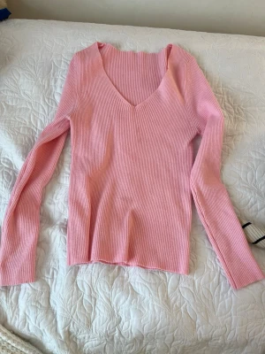 Rosa ribbad långärmad topp - Säljer en ljusrosa ribbad topp med v-ringning och långa ärmar. Toppen har en slim passform och är tillverkad i mjukt, stretchigt material som sitter snyggt på kroppen. Perfekt att styla med jeans eller kjol för en trendig look.