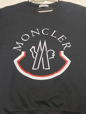 Svart Moncler tröja - Svart sweatshirt från Moncler i 100% bomull med stort vitt och rött logotryck på bröstet och klassisk Moncler–patch på ärmen. Rund hals och ribbade muddar ger en clean och stilren look. Perfekt för dig som gillar exklusiva streetwear–vibes. Den passar för både L och M dock är armarna lite korta och sizen är runt M och S