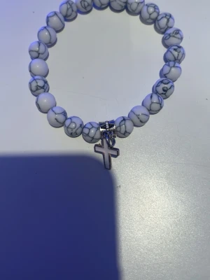 Vitt pärlarmband med korsberlock - Snyggt armband med vita pärlor som har grå marmorering och ett silverfärgat kors som hänge. Elastiskt band gör det enkelt att ta på och av. Perfekt accessoar för dig som gillar stilrena detaljer och vill addera något extra till din look.