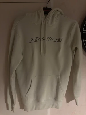 Vit Star Wars hoodie med Baby Yoda - Säljer en vit hoodie med Star Wars-tryck på bröstet och en stor Baby Yoda-illustration på ryggen. Hoodien har snörning i huvan och en stor magficka. Perfekt för dig som älskar Star Wars och vill ha en mysig och stilren hoodie.