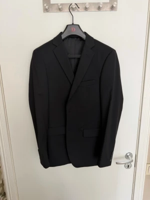 Svart klassisk kavaj - Stilren svart kavaj med två knappar framtill, smala slag och dubbla fickor med lock. Kavajen har en bröstficka och sprund baktill för extra rörelsefrihet. Perfekt för en dressad look och passar till skjorta eller t-shirt.