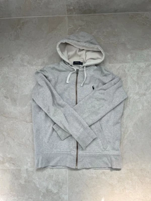 Grå hoodie från Polo Ralph Lauren - Snygg ljusgrå hoodie från Polo Ralph Lauren med dragkedja, vit dragsko och klassisk mörkblå logga broderad på bröstet. Hoodien har en mjuk insida och fickor framtill. Perfekt för en chill och stilren look. Skicket är extremt fint! Pris går att diskutera☺️
