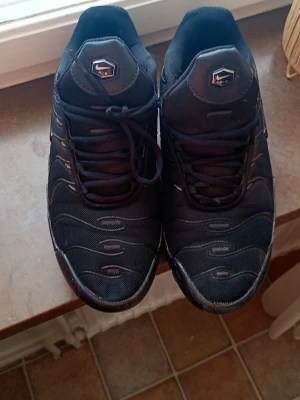 Svarta Nike Air Max Plus sneakers - Säljer ett par svarta Nike Air Max Plus sneakers med mesh- och syntetmaterial. Skorna har vågformade detaljer på ovandelen, klassisk Nike Air-logga på plösen och robust sula. Perfekta för dig som gillar streetwear och sportig stil.