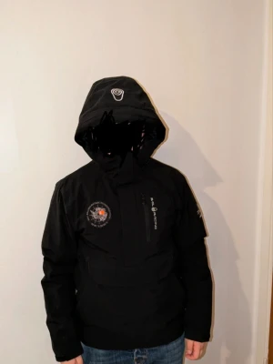 Svart vindjacka från Geographical Norway - Snygg svart vindjacka från Geographical Norway med huva och dragkedja framtill. Jackan har flera loggor och patchar på bröstet och ärmen, samt en praktisk bröstficka. Materialet är vindtätt och passar perfekt för blåsiga dagar.