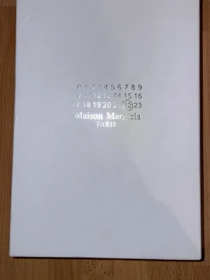 Maison Margiela svarta och grå sneakers - Snygga sneakers från Maison Margiela i svart läder med grå mockadetaljer och beige/brun sula. Klassisk låg modell med vita snören och stilren design som passar till det mesta. Perfekta för dig som gillar exklusiva och trendiga skor.