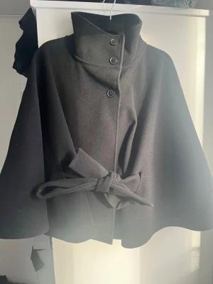 Svart poncho från Gina Tricot - Stilren svart cape-kappa från Gina Tricot med hög krage, knäppning med fyra knappar och ett brett knytband i midjan. Kappan har en vid, svepande siluett och är tillverkad i ett mjukt, tjockt tyg som ger en elegant look. ANVÄND 2-3 GÅNGER!🫶💕
