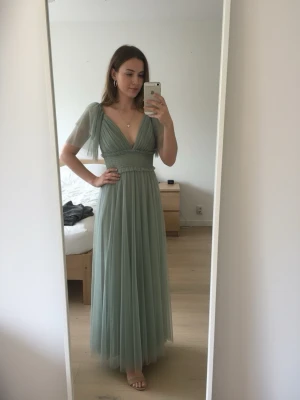 Ljusgrön långklänning med v-ringning - Elegant ljusgrön långklänning med djup v-ringning och korta, transparenta ärmar. Klänningen har ett markerat midjeband och ett flytande, lager på lager-tunt tyg i kjolen. Perfekt för sommarens speciella tillfällen.