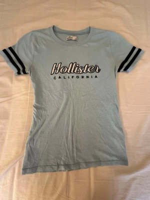 Ljusblå Hollister t-shirt med tryck - Snygg ljusblå t-shirt från Hollister med svart och vitt tryck på bröstet. T-shirten har korta ärmar med två svarta ränder på varje ärm och är gjord i mjuk bomull. Perfekt för en avslappnad och trendig look.