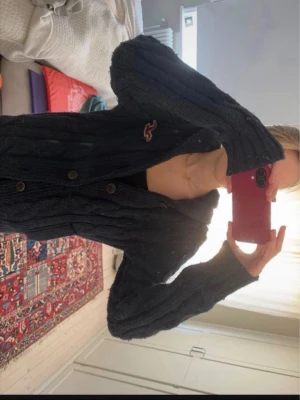  kofta Hollister - Säljer en mörkblå kabelstickad kofta från Hollister med stora knappar framtill och broderad logga på bröstet. Koftan har en mysig, tjock stickning och långa ärmar. Perfekt för lager på lager och chill dagar.