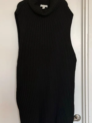 Svart stickad ärmlös polotröja - Säljer en svart stickad ärmlös polotröja med grov ribbstickning. Tröjan har hög polokrage och rak passform, perfekt att styla över en skjorta eller t-shirt. Materialet känns mjukt och tjockt, vilket gör den både trendig och bekväm. Lite nopprig som enkelt kan tas bort. 