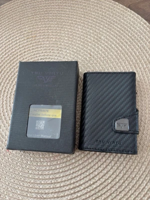 Svart plånbok från Tru Virtu - Snygg svart plånbok från Tru Virtu med coolt kolfibermönster och metallspänne med logotyp. Plånboken är kompakt och har en modern design som passar perfekt för kort och kontanter. Materialet är slitstarkt och ger en exklusiv känsla.