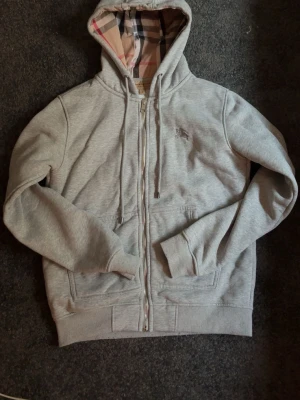 Grå zip hoodie från Burberry - Snygg grå hoodie med dragkedja från Burberry. Klassisk rutmönstrad huva i beige, svart och rött. Mjuk insida och två fickor framtill. Perfekt för dig som gillar stilrena och exklusiva plagg med ikoniska detaljer.