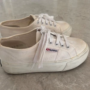 Vita Superga sneakers med platåsula - Säljer ett par vita Superga sneakers med platåsula och klassisk låg modell. Skorna har canvasöverdel, vita skosnören och metallöljetter. Superga-logga på sidan ger en clean och tidlös look. Perfekta för dig som gillar enkel och stilren streetwear.