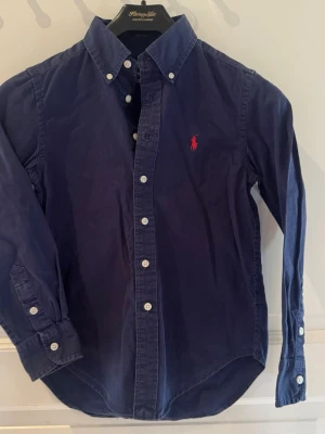 Marinblå skjorta från Ralph Lauren - Klassisk marinblå skjorta från Ralph Lauren med röd broderad logga på bröstet. Skjortan har lång ärm, knappar framtill och button-down krage. Perfekt till jeans eller chinos för en clean och stilren look. Sömmen har släppt lite nedtill på ryggen. Går lätt att laga.